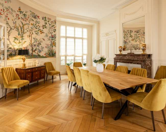 A spacious dining room features a long wooden table surrounded by yellow upholstered chairs, with floral wallpaper and natural light coming through large windows.