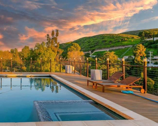 A modern swimming pool with wooden decking reflects the sunset sky, surrounded by lush greenery and hills in the background.