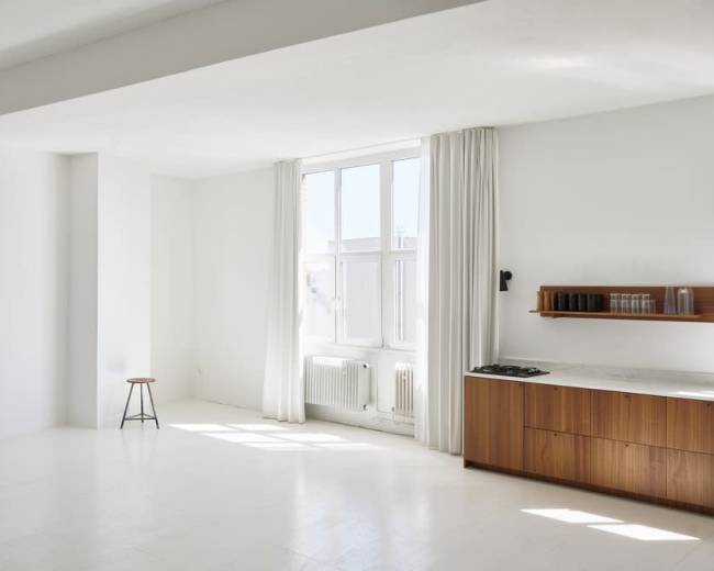 The image depicts a spacious, minimalistic room featuring a large window, white walls, a wooden countertop, and a stool.