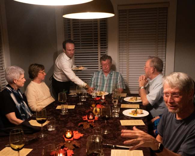 A group of eight people sit around a dining table adorned with candles and leaves as a server brings a dish to a guest.