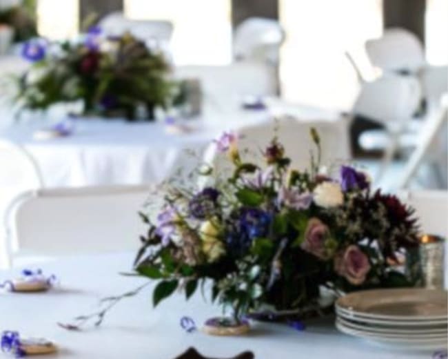 A white-tableclothed dining setup features several round tables adorned with floral centerpieces in shades of purple and green.