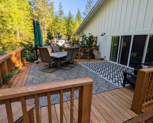 The image shows a wooden deck with patio furniture and a large outdoor rug, surrounded by trees.