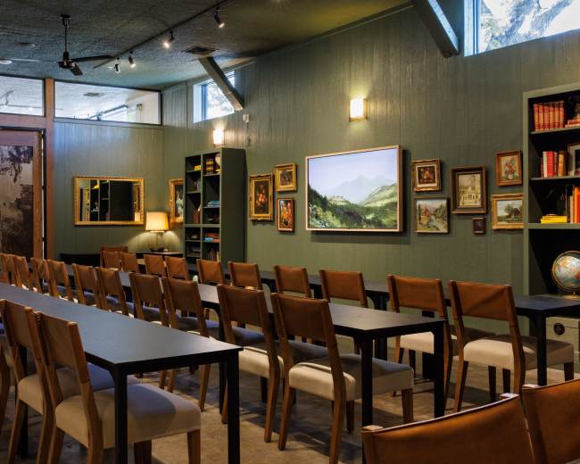 The image shows a long dining table surrounded by chairs in a room with green walls, lined with artwork and bookshelves.