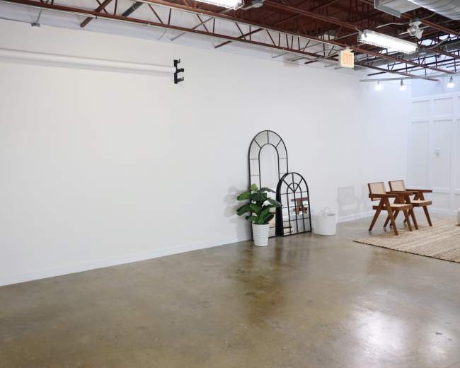 The image shows a spacious, empty room with a concrete floor, a few decorative elements including two arched mirrors and a potted plant, and a pair of wooden chairs on a woven rug.