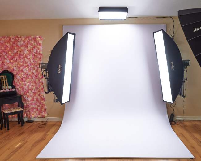 The image shows a photography studio setup featuring a white backdrop, two softbox lights on either side, and a patterned fabric drape in the background.