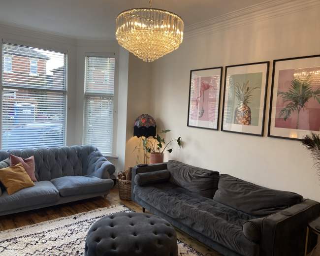 The living room features two plush sofas, a round ottoman, a crystal chandelier, and framed art depicting plants on the walls.