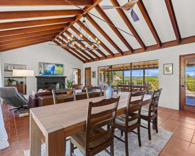 The image shows a spacious dining area with a wooden table surrounded by chairs, featuring exposed beam ceilings and large windows that provide a view of the outdoors.