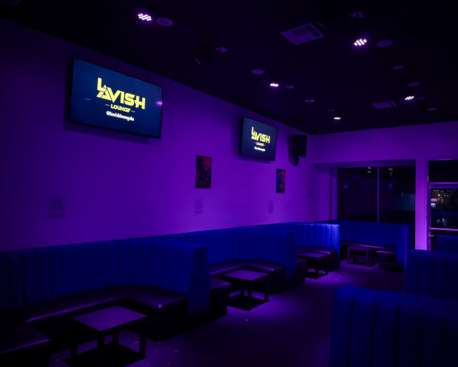The image shows a dimly lit lounge with purple lighting, featuring blue booths and two TV screens displaying the word "LAVISH."