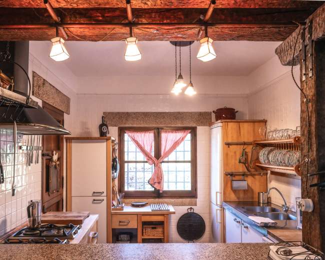 A kitchen with wooden beams, a window with a red checkered curtain, and various cooking appliances and utensils neatly arranged.