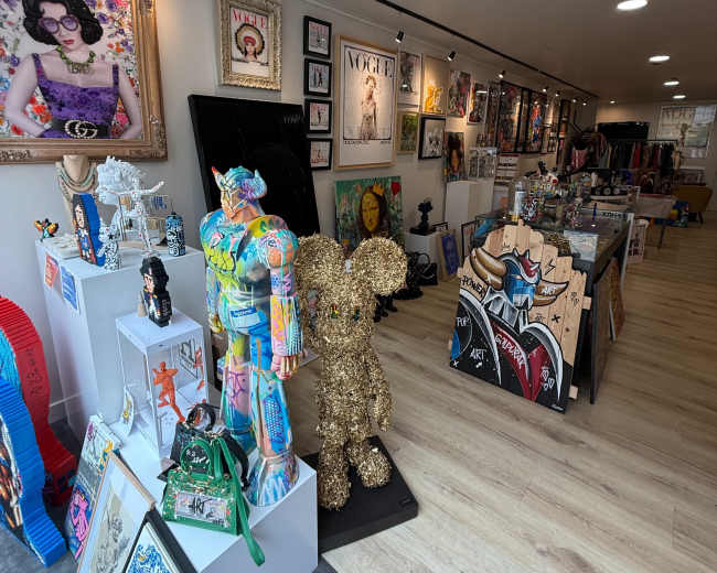 The image shows a colorful art gallery featuring various artwork and sculptures, with displays along the walls and several pieces arranged on stands and tables.