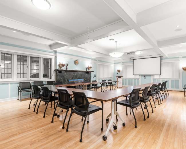 The image shows a spacious meeting room with a long table set up in the center, surrounded by chairs, and equipped with a screen at one end.
