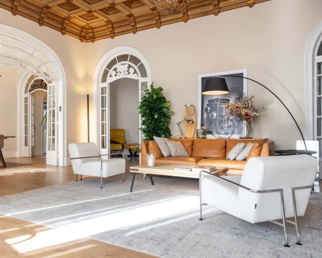 The image shows a stylish living area with a leather sofa, modern chairs, a coffee table, and large windows illuminating the space.