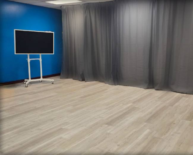 The image shows a room with grey curtains draping on one side and a large screen on a stand set against a blue wall.