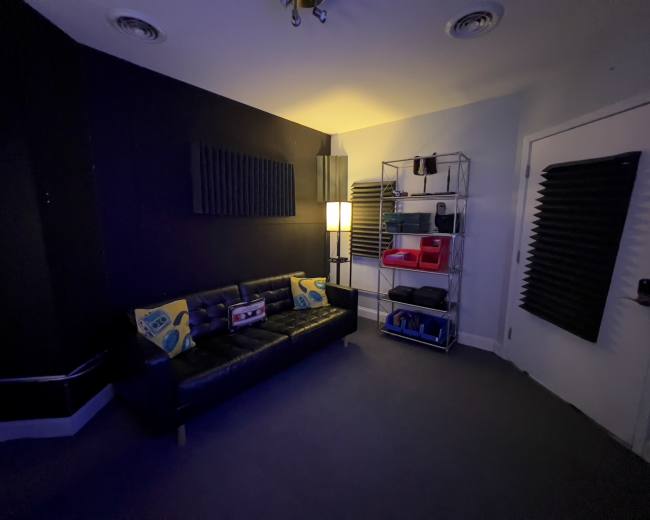The image shows a dark room with a black wall, a couch with patterned cushions, a lamp, and a wire shelf containing various items.