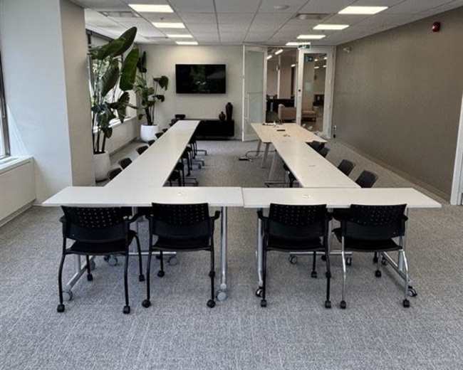 The image shows a modern conference room with several long tables arranged in a U-shape, accompanied by black rolling chairs and large windows.