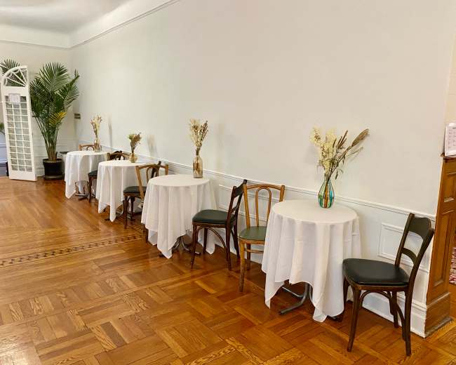 A row of tables covered with white tablecloths and adorned with vases of dried flowers is lined against a wall in a room with wooden flooring.