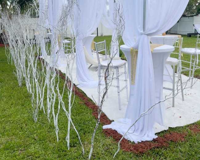 A white tent with sheer curtains is set up outside on a grassy area, surrounded by bare twigs arranged decoratively.