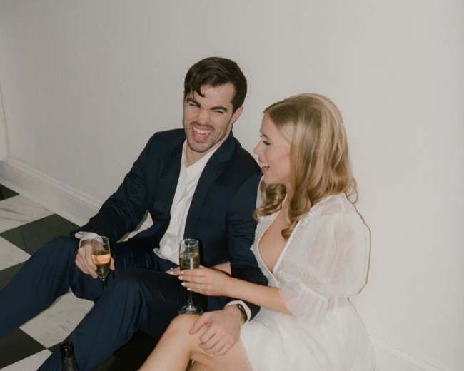 A couple sits on a checkered floor, smiling and holding champagne glasses while wearing formal attire.