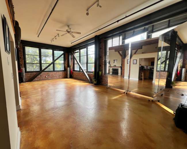The image shows a spacious, industrial-style room with large windows, exposed brick walls, and polished concrete flooring.