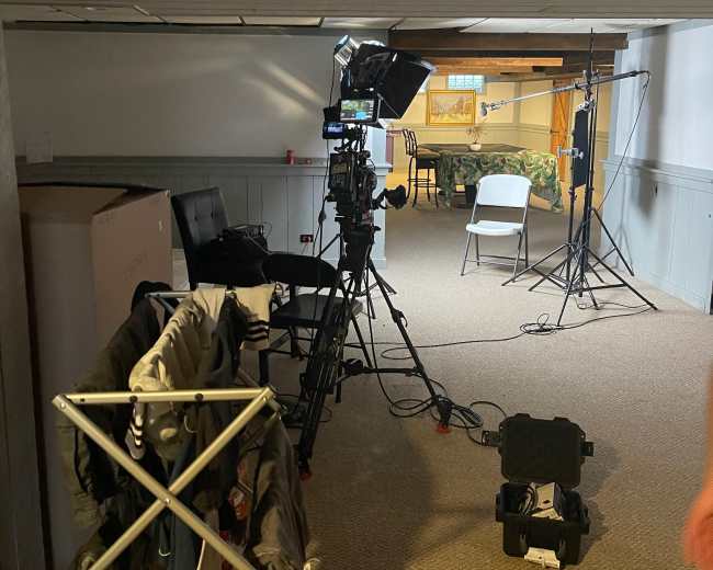 A filming setup in a room, with a camera, lights, and empty chairs arranged for a shoot.