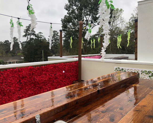 The image shows a wooden table on a rooftop adorned with decorative flowers and greenery hanging from overhead lines, with a cloudy sky in the background.