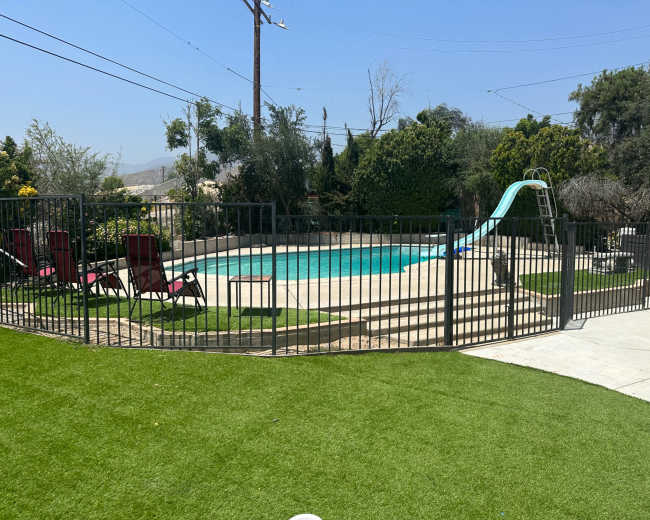 The image shows a fenced-in swimming pool surrounded by green grass and several lounge chairs.