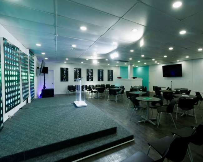 The image shows a modern event space with a stage, rows of black chairs, and a bar area in the background.