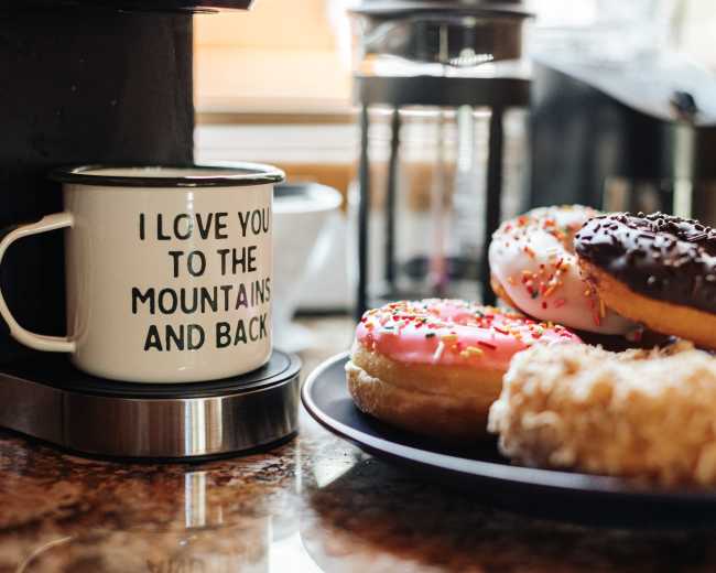 A coffee mug with the phrase "I love you to the mountains and back" sits next to a platter of assorted donuts on a kitchen countertop.
