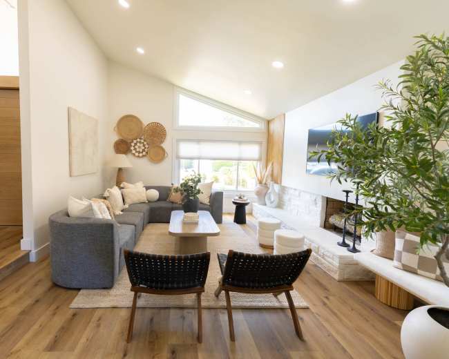 A modern living room features a gray sectional sofa, two black chairs, a coffee table, and a large window with natural light, accented by decorative wall art and greenery.