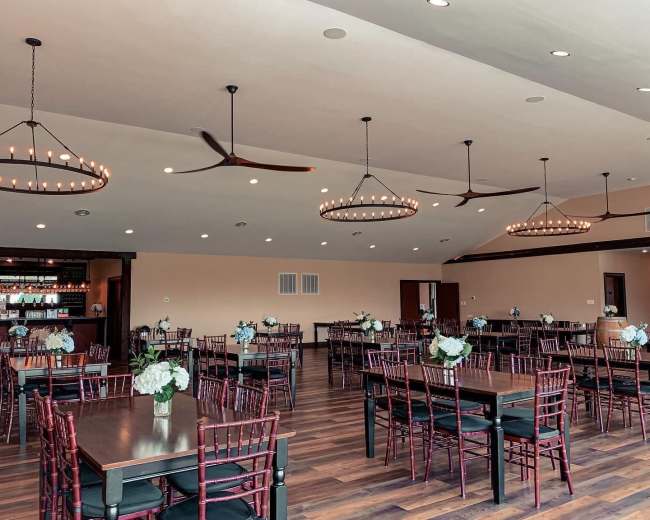 The image shows a spacious dining area with multiple tables arranged neatly, each adorned with flower centerpieces, and featuring modern chandeliers and ceiling fans.