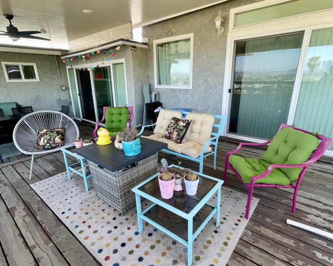 The image shows a patio furnished with colorful chairs, a coffee table, and potted plants on a wooden deck under a shaded roof.
