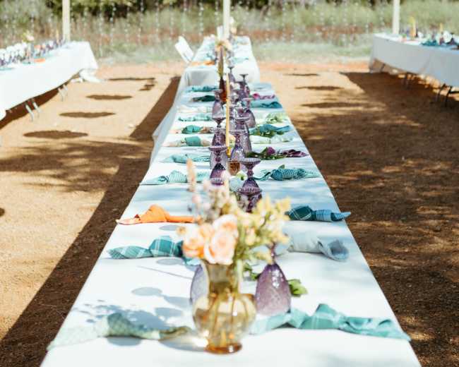 A long dining table is set outdoors, adorned with white tablecloths, colorful napkins, and vases of flowers, while oversized white lanterns hang above.
