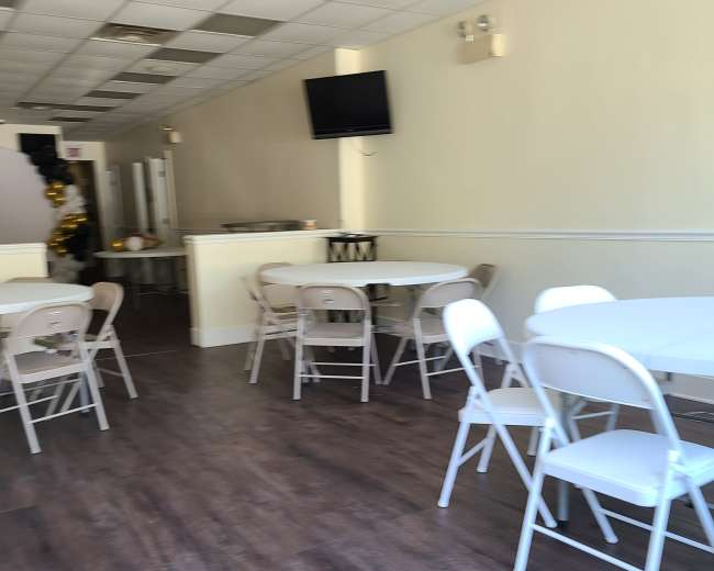 The image shows a spacious interior with several white folding tables and chairs arranged on a wooden floor, along with a television mounted on the wall.