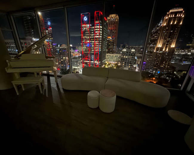 The image shows a modern interior with a curved white sofa and a piano, overlooking a city skyline lit up at night.
