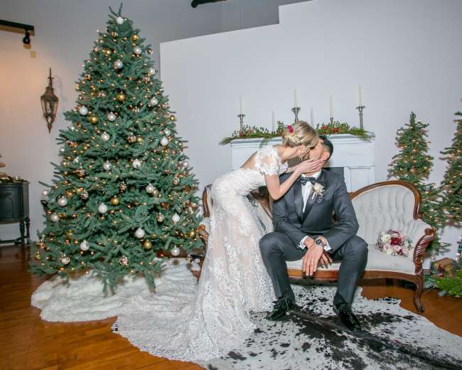 A bride kisses a groom while sitting on a vintage sofa, with a decorated Christmas tree and smaller evergreen trees in the background.
