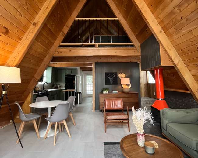 The image shows the interior of a spacious A-frame cabin featuring wooden beams, a dining area with modern chairs, and a cozy living space with a fireplace.