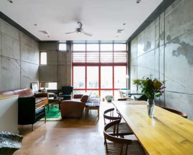 A modern living space with concrete walls, a wooden dining table, and large windows allowing natural light to enter.