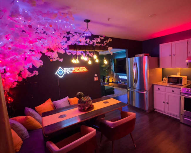 A modern kitchen features a dining area with a table, surrounded by colorful lighting and decorative elements like artificial flowers.