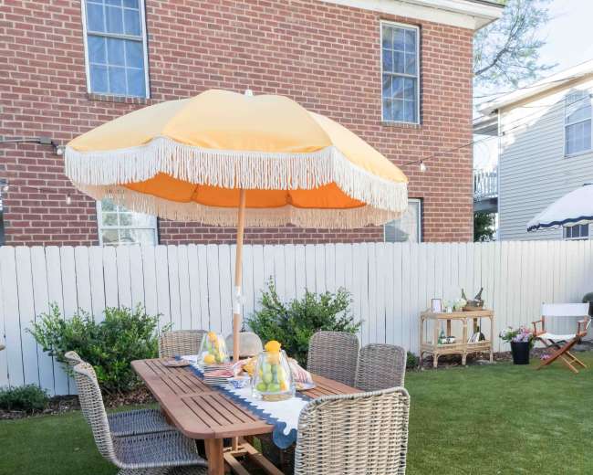 A patio set with four wicker chairs and a wooden table is located on a grassy area, shaded by a large orange umbrella under a blue sky.