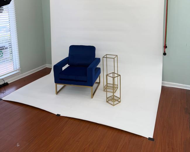 The image shows a photography studio with a blue chair and a gold side table against a white backdrop.