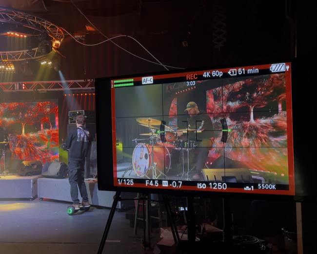 A musician performs on stage with a drum set and vibrant visual displays in the background, while a camera monitor captures the scene.