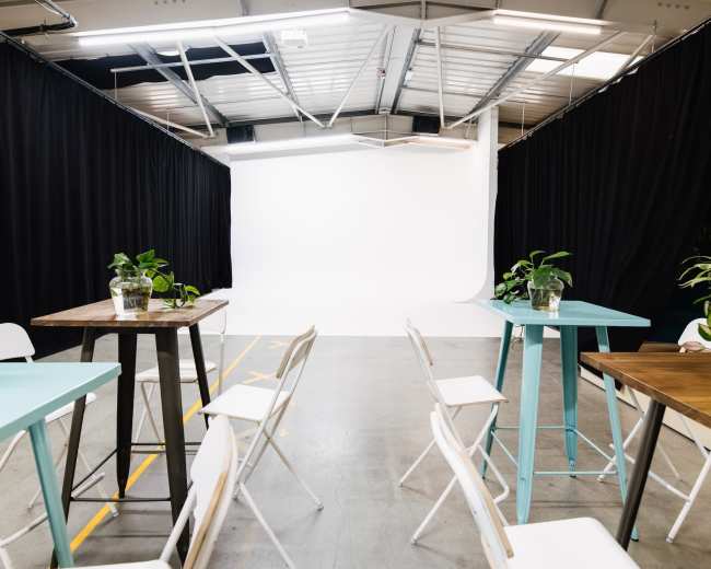 A spacious studio features several tables and chairs arranged in rows, leading to a plain white backdrop at the far end.