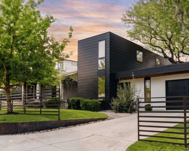 The image shows a modern two-story house with a black exterior located on a landscaped property featuring a curved driveway and surrounding greenery.
