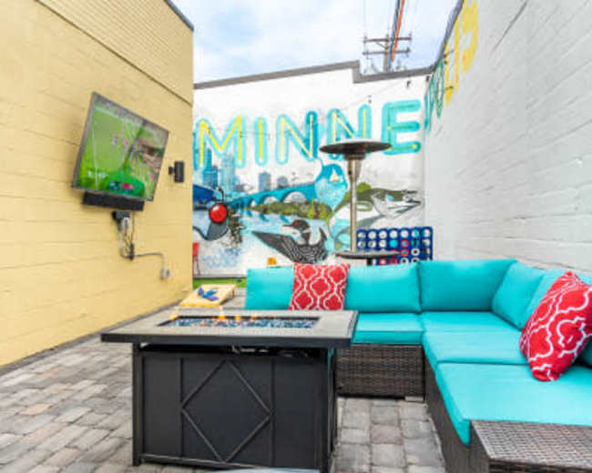 A small outdoor seating area featuring a blue sectional sofa with red accent pillows, a fire pit table, and a wall mural depicting a cityscape.