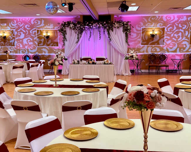 The banquet hall features elegantly set tables with gold chargers and white tablecloths, adorned with red sashes, under decorative lighting and floral arrangements.