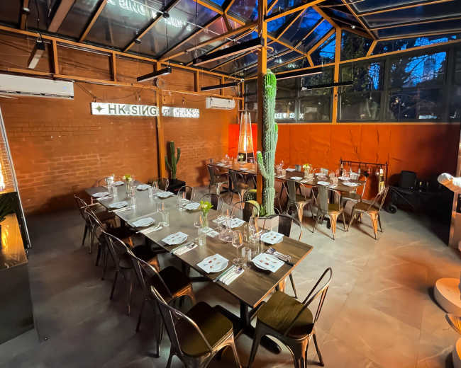 A well-arranged dining area features tables set with cutlery and glassware under a glass ceiling, illuminated by ambient lighting and decorative plants.