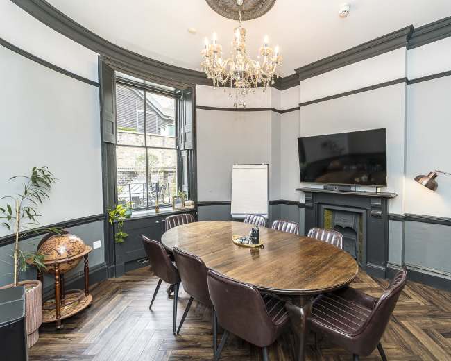 The image shows a well-appointed meeting room featuring a round wooden table surrounded by brown leather chairs, a large wall-mounted TV, and a chandelier hanging from the ceiling, with windows allowing natural light.