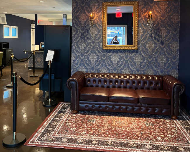 The image shows a lobby area featuring a brown leather chesterfield sofa, ornate wallpaper, a decorative mirror, and a patterned rug, with a stanchion barrier visible in the foreground.