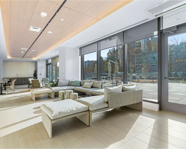 The image shows a modern lounge area with a sectional sofa, coffee table, and large windows allowing natural light to fill the space.