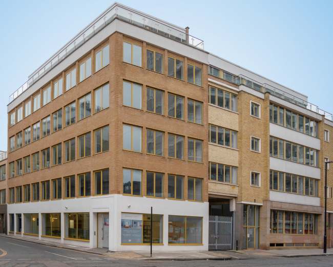 A four-story brick building with large windows stands on a street corner, featuring a modern design and bordered by a paved sidewalk.